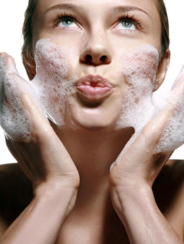 Close-up of a fair-skinned woman with blue eyes, puckered lips, and soapy white foam on her face and hands, set against a plain white backdrop.