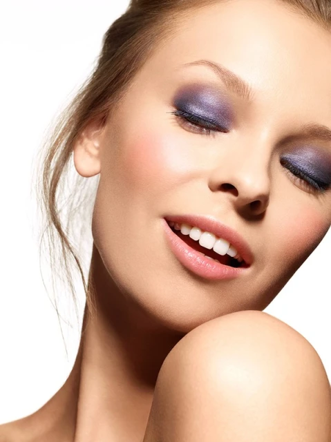 Close-up portrait of a light-skinned adult woman with blue eyeshadow, pink lipstick, and bare shoulders against a white backdrop.