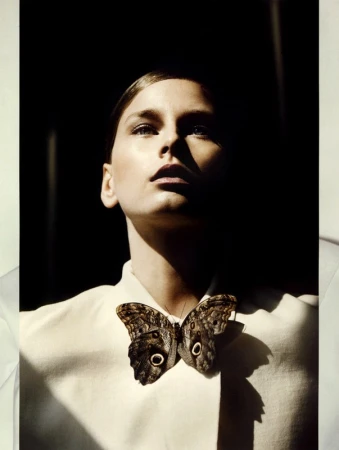 Close-up portrait of a woman wearing a white collared garment with a large patterned butterfly brooch on her chest under dramatic lighting.
