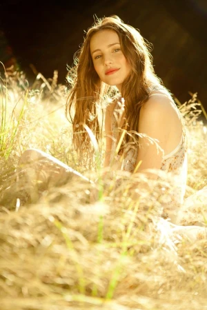 Young woman with long brown hair seated outdoors amidst tall golden grass illuminated by warm sunlight.