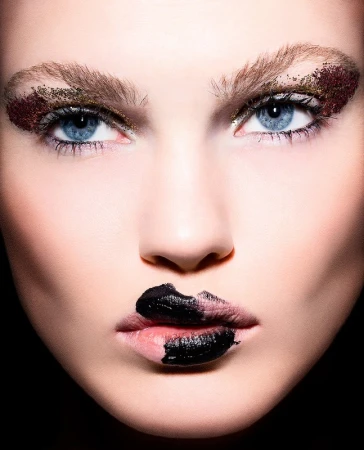 Close-up beauty portrait of a woman with blue eyes, glitter eye makeup, and bold textured black lipstick by Jenna Menard
