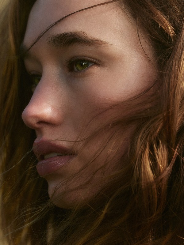 Close-up portrait of a young adult female with long brown hair and green eyes, in an outdoor setting with diffused lighting.