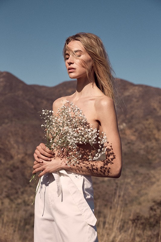 Blonde woman with bare shoulders poses outdoors with a bouquet of white flowers in front of brown mountains under a blue sky.