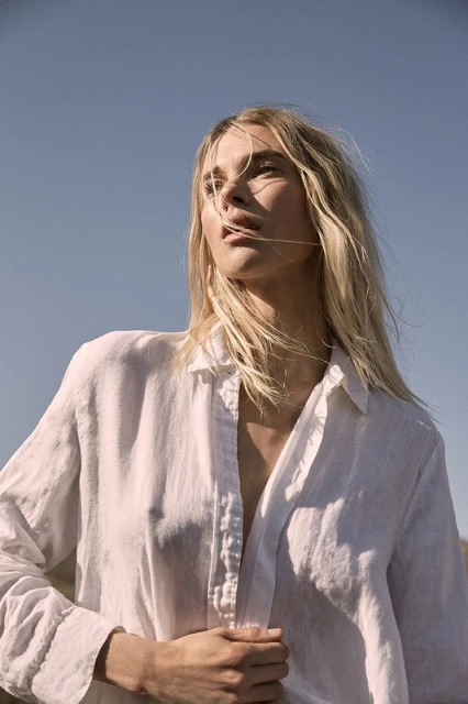 Woman with blonde hair wearing a white linen shirt poses in an outdoor setting under a blue sky. Fashion photography, natural light.
