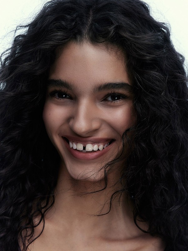 Close-up portrait of a young woman with dark curly hair, smiling broadly to reveal a gap in her front teeth, against a light backdrop.