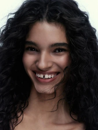 Close-up portrait of a smiling young woman with dark curly hair and visible gap between front teeth against a neutral backdrop.