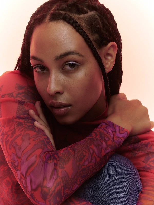 Portrait of a young woman with braided hair in a red patterned top, seated indoors with arms crossed against a soft-lit background.