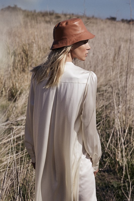Woman in nature wearing a leather bucket hat and long sleeve shirt. Outdoor setting with tall grass and a blue sky. Fashion shoot.