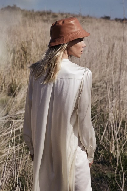Woman in nature wearing a leather bucket hat and long sleeve shirt. Outdoor setting with tall grass and a blue sky. Fashion shoot.