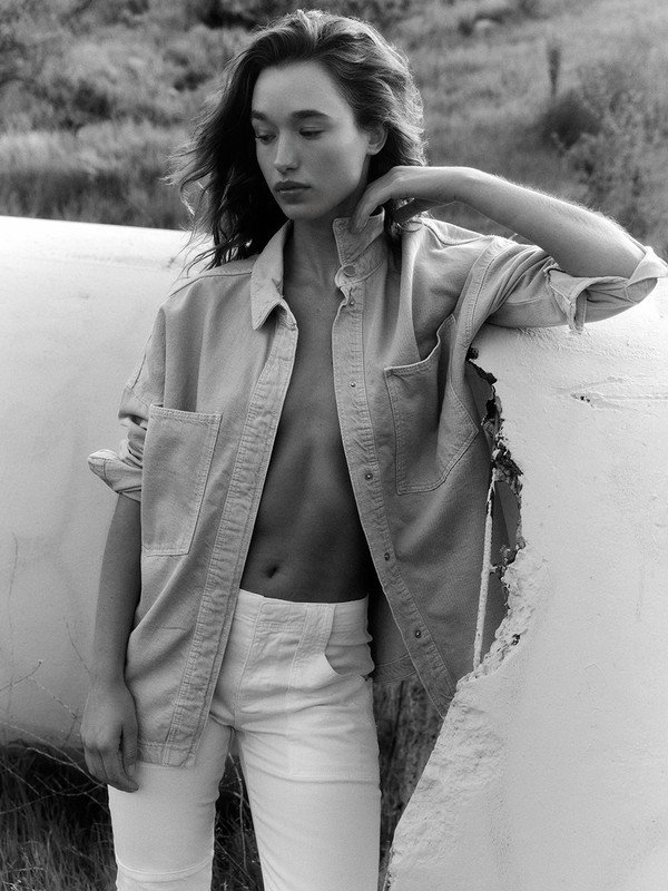 Monochrome portrait of a young woman in an unbuttoned shirt and white pants leaning on a weathered structure outdoors in a field.