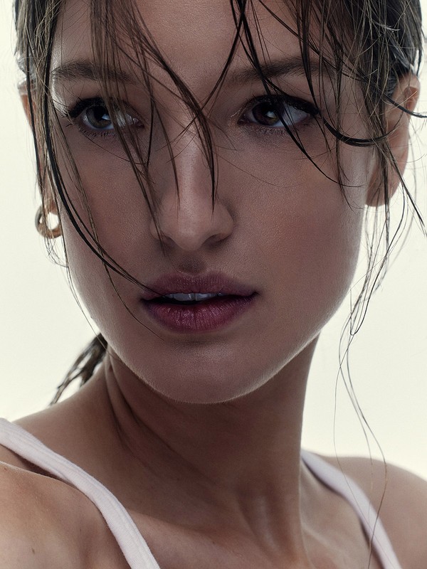 Close-up portrait of a woman with wet hair, fair skin, and a white tank top, set against a neutral background in soft lighting.