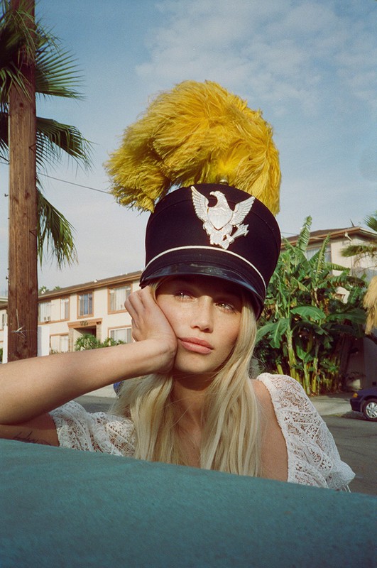 Portrait of a blonde woman in a marching band hat with a feather, resting her head on her hand, with palm trees in background.