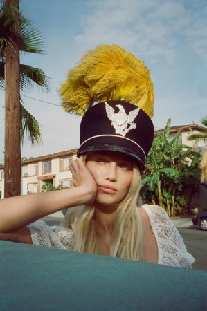 Portrait of a blonde woman in a marching band hat with a feather, resting her head on her hand, with palm trees in background.