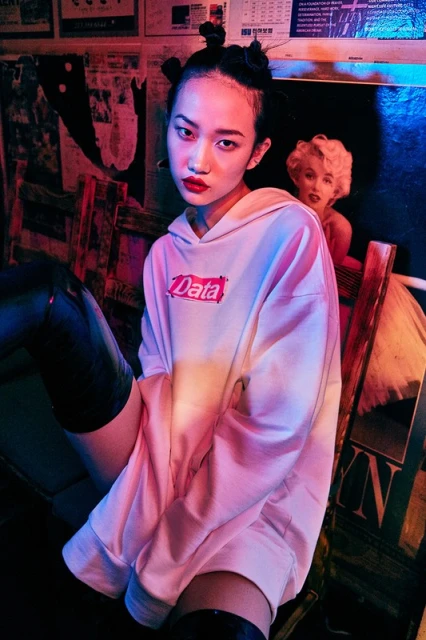 A young Asian woman in a white hoodie with the word 'Data' poses in front of a poster of Marilyn Monroe, illuminated by pink and blue lighting.