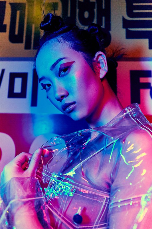 Woman with creative makeup and hair wearing transparent vinyl jacket, studio shot with blue and pink lighting. Beauty editorial.