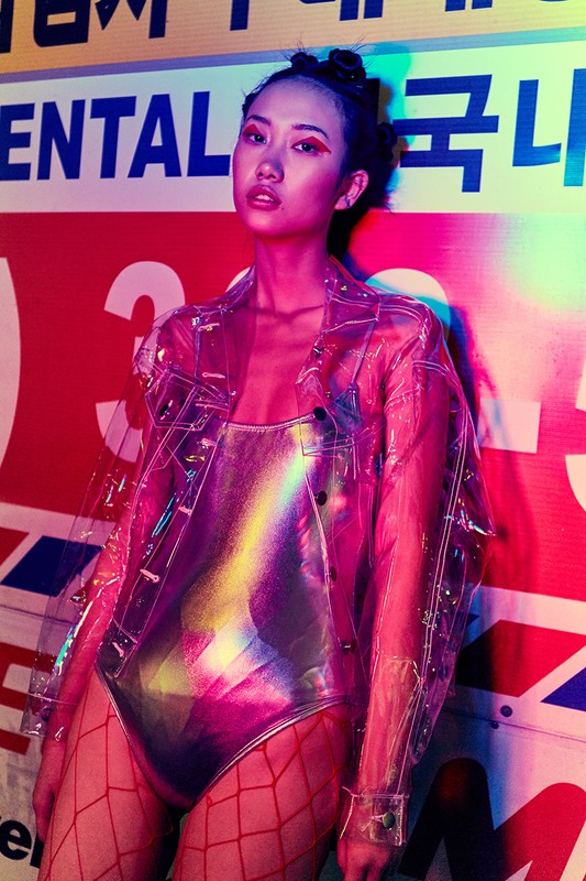 Woman in iridescent bodysuit and clear jacket poses against a red, white, and blue wall with Korean lettering and neon lighting.