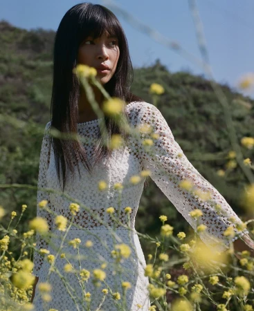 A woman in a white lace dress posing outdoors among yellow wildflowers with natural lighting.