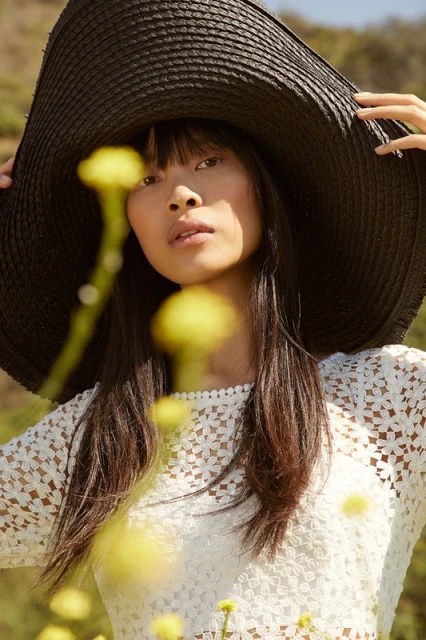 Close-up of a young Asian woman in a large black hat and white lace top, framed by blurred yellow wildflowers, against an outdoor background.