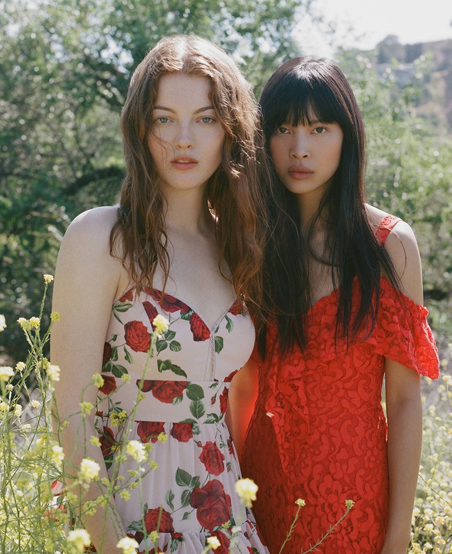 Two women pose in a sunlit meadow; one in a rose-patterned dress, the other in red lace, amidst wildflowers and natural outdoor scenery.