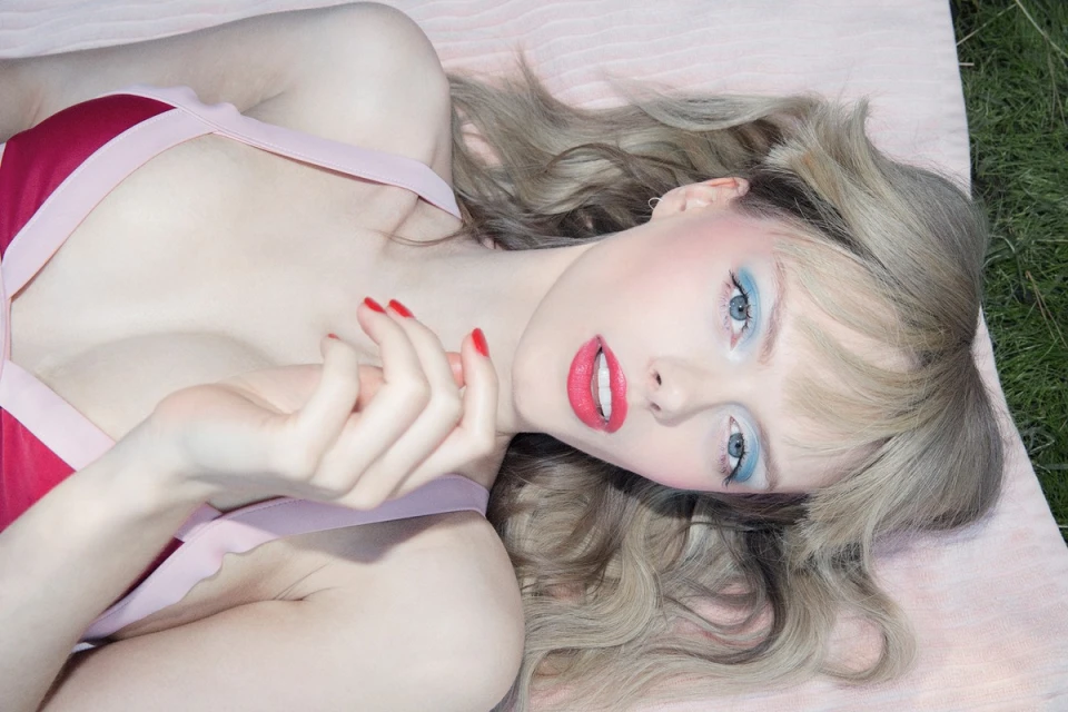 Close-up of fair-skinned woman with wavy blonde hair, red lipstick and red nails lying outdoors on a light pink blanket in green grass.