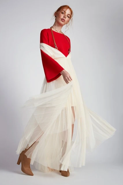A red-haired woman wearing a red top and ivory tulle skirt poses in a studio, set against a clean, neutral backdrop.