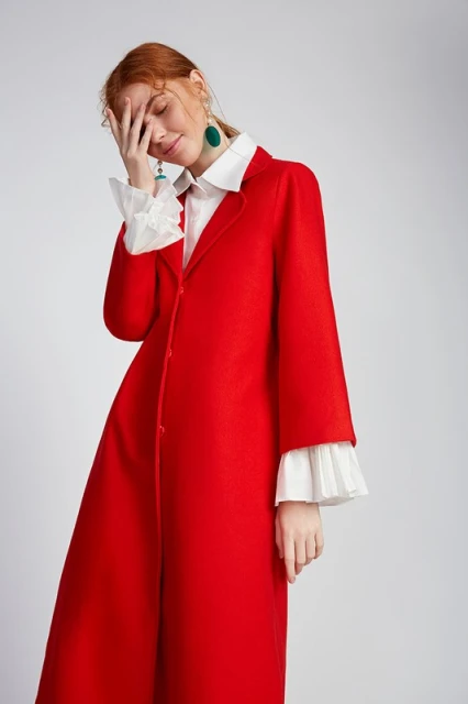 A red-haired woman wearing a red coat, white blouse, and teal earrings raises her hand to her face in a fashion shot against a grey backdrop.