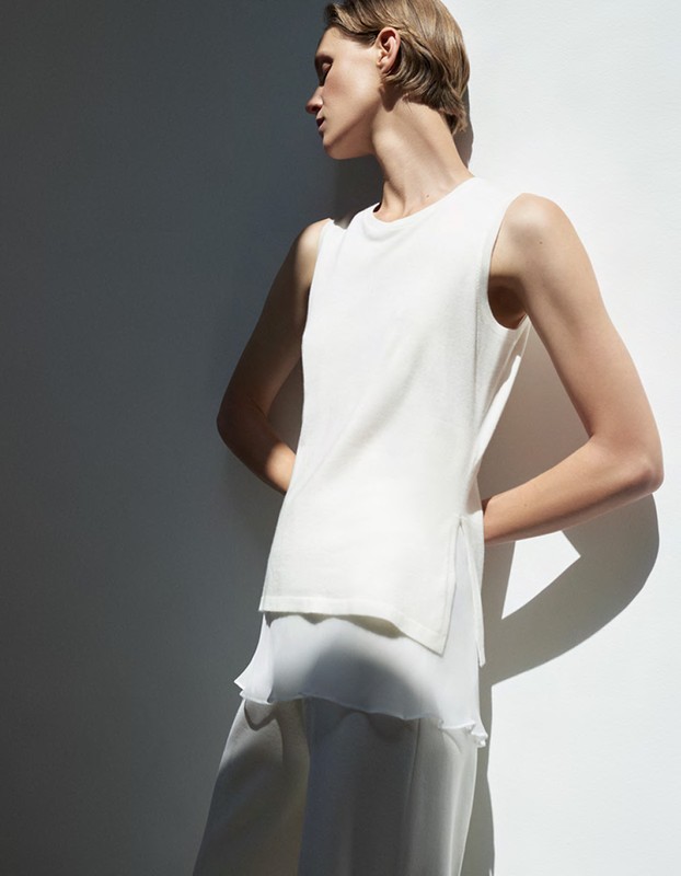 Woman in a sleeveless white top and chiffon pants stands against a white wall with shadows, showcasing a minimalist fashion style.