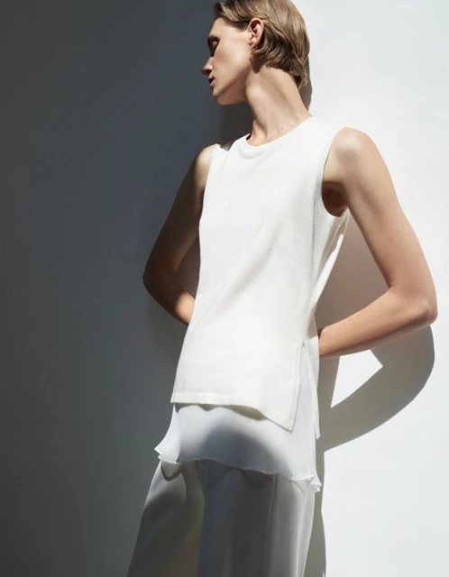 Woman in a sleeveless white top and chiffon pants stands against a white wall with shadows, showcasing a minimalist fashion style.