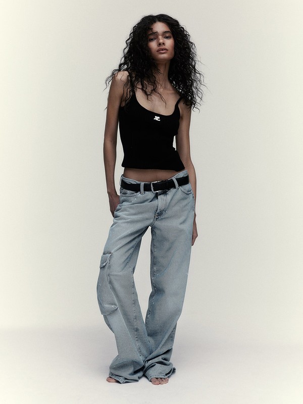 Full-length studio shot of a slender female model posing in a black tank top, wide-leg jeans, and a black belt, with soft studio lighting.
