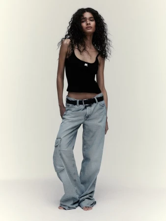 Full-length studio shot of a slender female model posing in a black tank top, wide-leg jeans, and a black belt, with soft studio lighting.