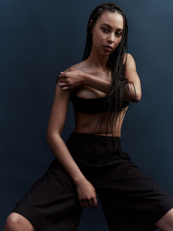 Portrait of a young adult woman with braids wearing a black top and pants, posing against a dark blue background in a photo studio.