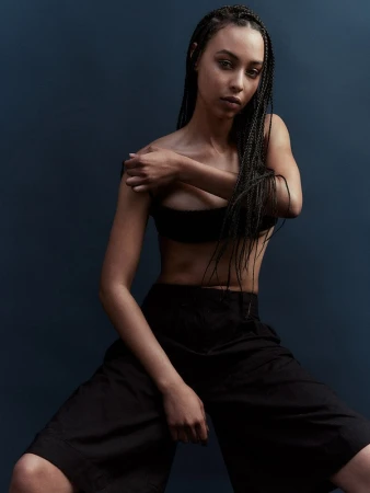 Portrait of a young adult woman with braids wearing a black top and pants, posing against a dark blue background in a photo studio.