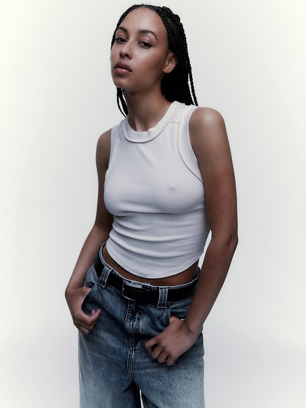 A young woman with braided hair poses in a light tank top and high-waisted jeans against a neutral background in a medium studio shot.
