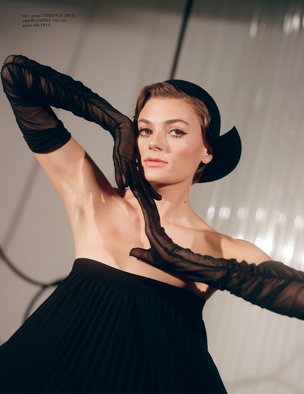 Model in black strapless dress and sheer long gloves posing with sculptural hat against a light background