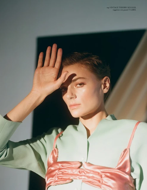 Portrait of a young woman with short hair wearing a light green jacket and pink satin detail, posing with her hand up to the sun.