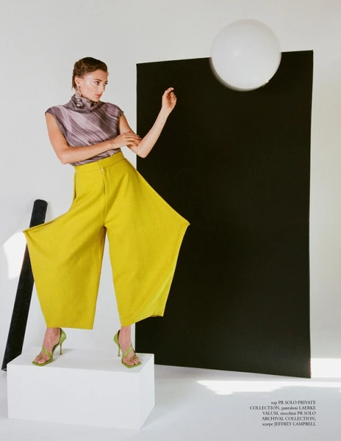 A fashion model poses in a photo studio wearing a purple top and wide-legged chartreuse pants, complemented by matching green high-heeled shoes.