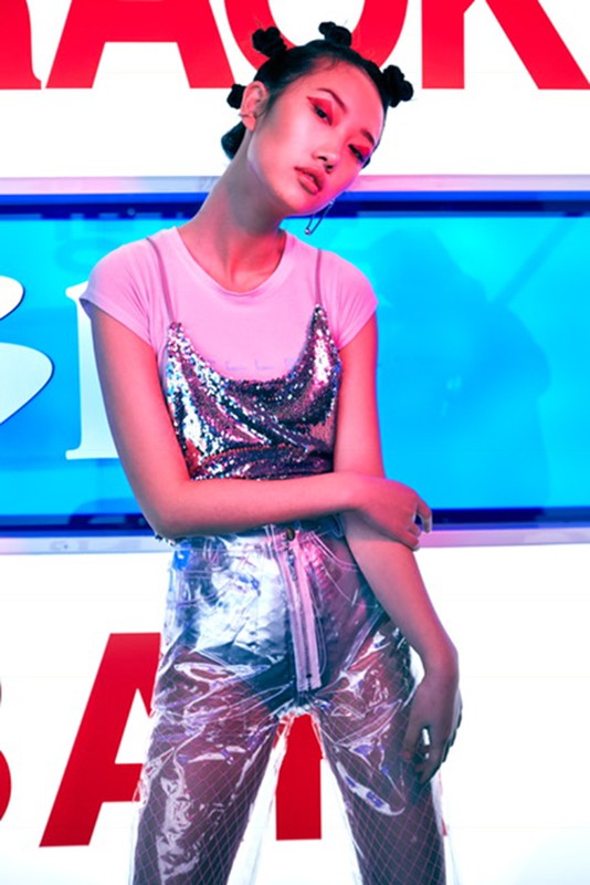 A young Asian woman poses in a pink shirt and clear vinyl pants in a studio with neon lights and bold lettered background signs.