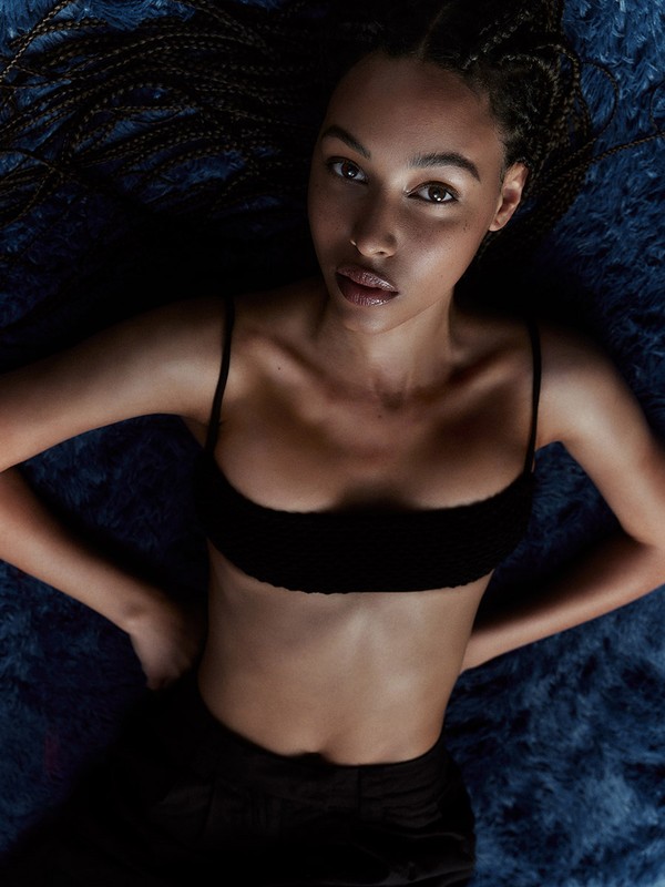 Portrait of a young adult female model with braided hair, wearing a black bralette, laying on a blue textured background, and looking at the camera.