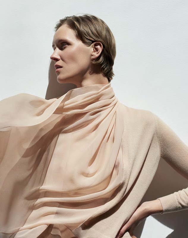 A woman with short blond hair poses in a beige sweater with a draped sheer scarf, standing against a white wall with natural light.