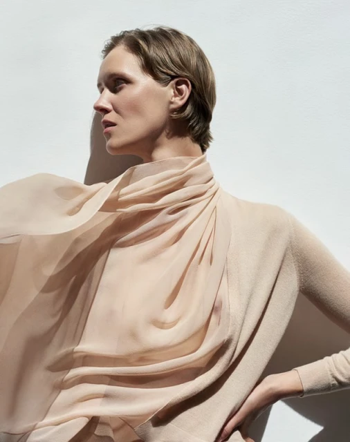 A woman with short blond hair poses in a beige sweater with a draped sheer scarf, standing against a white wall with natural light.