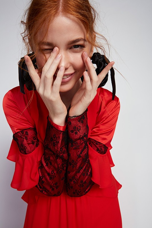 Red-haired woman in a red dress with black lace sleeves hiding her face behind hands, styled with hair scrunchies.