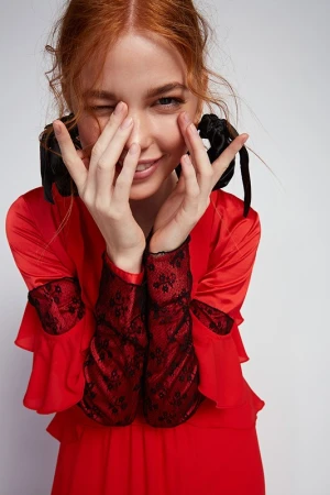 Red-haired woman in a red dress with black lace sleeves hiding her face behind hands, styled with hair scrunchies.