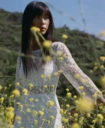 A woman in a white lace dress posing outdoors among yellow wildflowers with natural lighting.