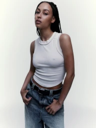 Woman with braided dark hair wearing a white tank top and blue denim jeans against a seamless white background.