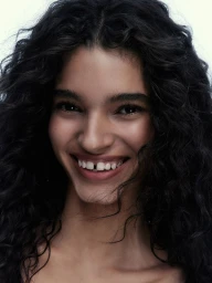 Close-up portrait of a smiling young woman with dark curly hair and visible gap between front teeth against a neutral backdrop.