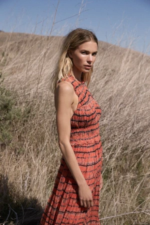 Blonde female model in an orange patterned dress standing outdoors among dry tall grass under a clear blue sky