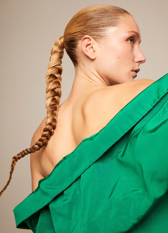 Woman with long braid and bare back draped in green fabric in a studio portrait with a neutral background. Beauty and fashion concept.