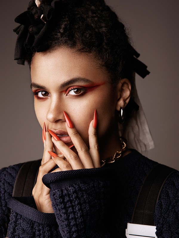 Close-up portrait of a woman with dark hair, wearing a sweater and gold chain, posing with hands near face, red makeup, and long orange nails.