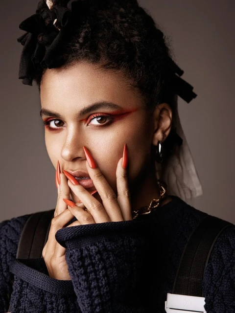 Close-up portrait of a woman with dark hair, wearing a sweater and gold chain, posing with hands near face, red makeup, and long orange nails.