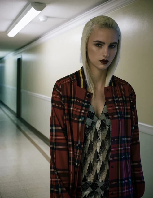 Indoor portrait of a fair-skinned young adult woman with blonde hair, wearing a plaid jacket and dress, standing in a hallway.